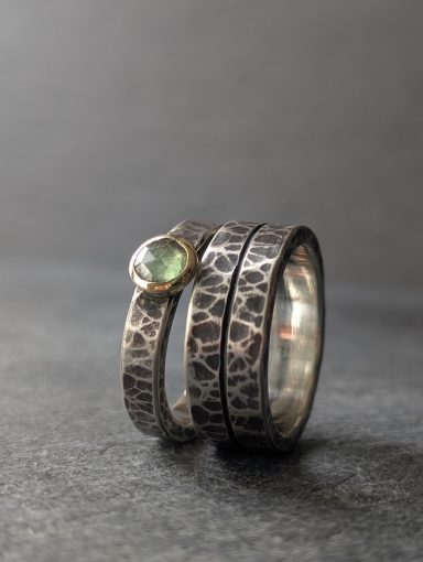 Verlobungsringe aus Silber mit dunkler Textur auf grauem Untergrund. Der Damenring hat einen grünen Kyanit in Goldfassung