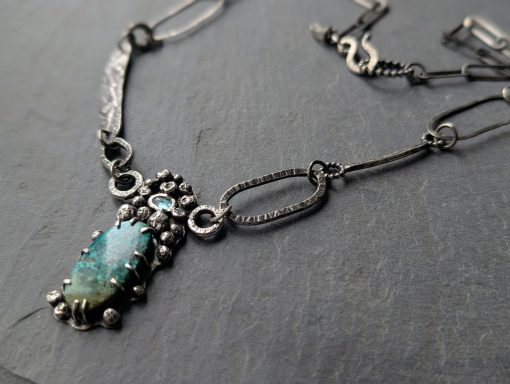 Eine massive Silberkette mit einem Mittelteil in den ein Türkis und ein blauer Apatit eingefasst ist, liegt auf einem grauen Stein